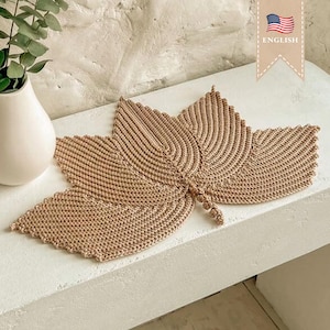 May include: A beige, crocheted leaf-shaped placemat. The placemat is made of a thick, textured yarn and features detailed ribbing to mimic the veins of a leaf. The leaf is displayed on a white surface, with a white vase and greenery in the background.