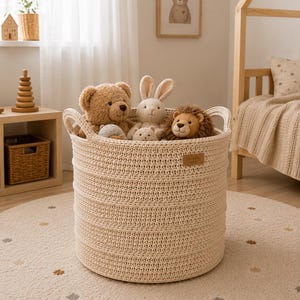 Puede incluir: Una cesta de almacenamiento redonda de tejido color crema con dos asas integradas, llena de varios peluches, incluyendo un oso marr&oacute;n, un conejo de color tostado claro y un le&oacute;n. Una peque&ntilde;a etiqueta rectangular de piel sint&eacute;tica est&aacute; fijada en el lateral.