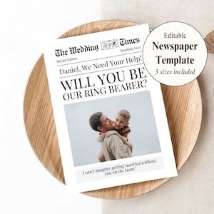 May include: A white card in the style of a newspaper with a black border and text that reads "The Wedding Times, Special Edition, Breaking News, Daniel, We Need Your Help! Will You Be Our Ring Bearer? I can't imagine getting married without you on the team!" There is a photo of two men, one with his arm around the other, smiling. The card is on a wooden surface.