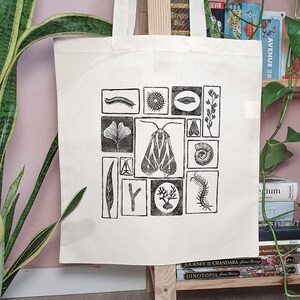May include: Cream-colored tote bag featuring a black and white linocut print of various insects, plants, and other natural elements. The design includes a moth, a leaf, a centipede, and other botanical and zoological illustrations. The bag is made of a sturdy fabric.