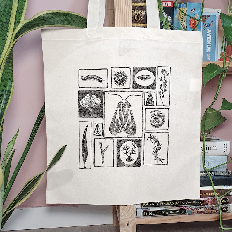 May include: Cream-colored tote bag featuring a black and white linocut print of various insects, plants, and other natural elements. The design includes a moth, a leaf, a centipede, and other botanical and zoological illustrations. The bag is made of a sturdy fabric.