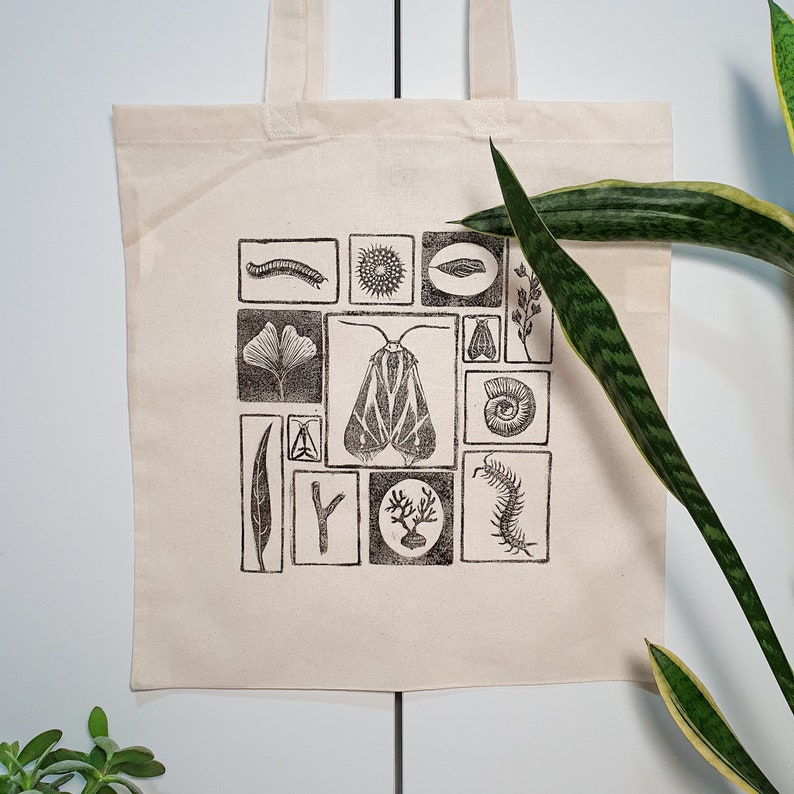 May include: Cream-colored tote bag featuring a black and white block print design of various insects, leaves, and other natural elements. The design is arranged in a grid pattern. The bag has two handles and is made of a sturdy fabric.