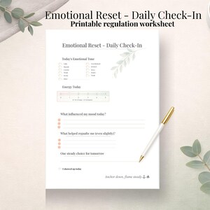 May include: A white printable worksheet titled "Emotional Reset - Daily Check-In" with sections for emotional tone, energy levels, and mood influences. A gold-tipped pen and a cup of coffee are on the table.
