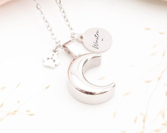 May include: A silver-toned necklace featuring a crescent moon pendant, a small circular charm engraved with the name Winston, and a tiny paw print charm. The metal chain is delicate and polished, reflecting light across the smooth surfaces.