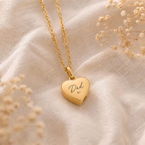 May include: A gold-toned heart-shaped pendant necklace featuring a delicate chain and a smooth, polished surface. The centre of the heart is engraved with the word Dad in a cursive script, followed by a small x symbol, creating a simple design.