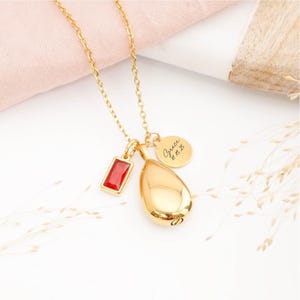Gold teardrop urn necklace with July birthstone and engraved memorial tag on wooden table