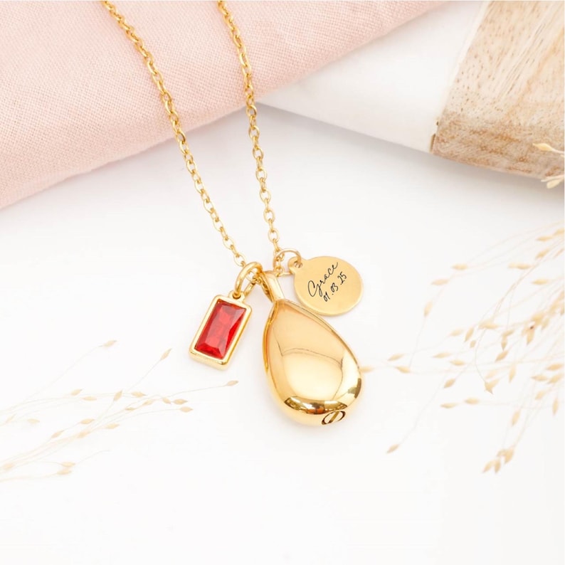 Gold teardrop urn necklace with July birthstone and engraved memorial tag on wooden table