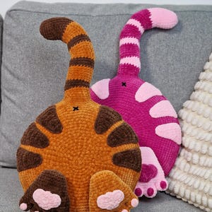May include: Two handmade cat butt pillows. One is orange with brown stripes and paw pads, the other is pink with white spots and paw pads. Both have a tail that curves upwards. They are placed on a gray couch.
