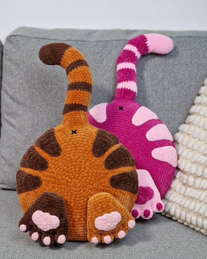 May include: Two handmade cat butt pillows. One is orange with brown stripes and paw pads, the other is pink with white spots and paw pads. Both have a tail that curves upwards. They are placed on a gray couch.