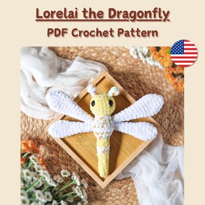Pode incluir: Uma libélula de crochê amarela e branca, chamada Lorelai, em uma bandeja de madeira. A libélula tem asas brancas e um corpo estampado. O texto na imagem diz "Lorelai the Dragonfly PDF Crochet Pattern" e "No sewing required."
