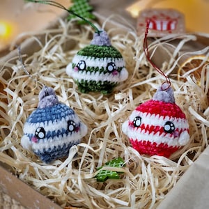 Op de afbeelding: Drie handgemaakte gehaakte kerstballen in een decoratieve doos. De ornamenten zijn rood, groen en blauw, met witte strepen en cartoongezichten. Elk heeft een zilveren of groen touwtje om op te hangen. De doos is gevuld met stro.