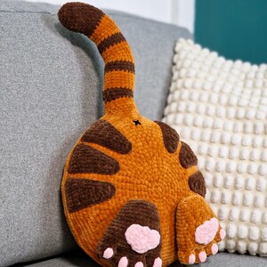 May include: A handmade orange and brown cat butt pillow with a striped tail and brown spots. The pillow features two brown paws with pink paw pads. The plush pillow is designed to look like a cat's rear end.