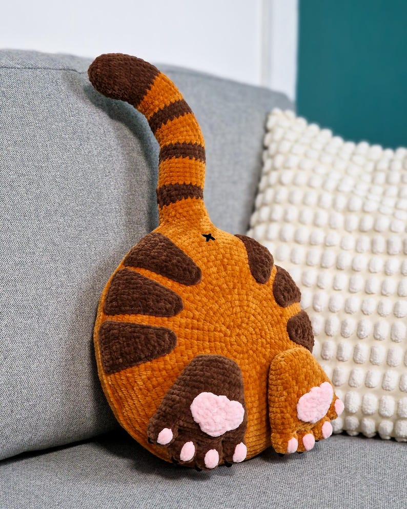 May include: A handmade orange and brown cat butt pillow with a striped tail and brown spots. The pillow features two brown paws with pink paw pads. The plush pillow is designed to look like a cat's rear end.
