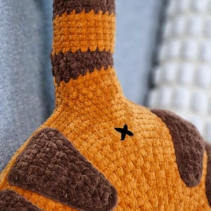 May include: Close-up of a plush toy tail in shades of orange and brown. The tail has brown stripes and a small black cross-stitch detail. The plush material appears soft and textured, suggesting a handmade item.