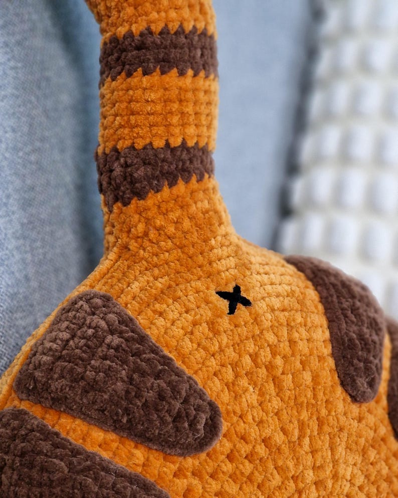 May include: Close-up of a plush toy tail in shades of orange and brown. The tail has brown stripes and a small black cross-stitch detail. The plush material appears soft and textured, suggesting a handmade item.