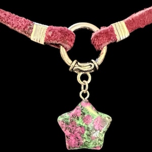 Könnte beinhalten: Eine bordeauxfarbene Samt-Halskette mit einem silberfarbenen Ringverschluss. Ein sternförmiger Anhänger mit rosa und grünem Stein hängt an dem Ring. Der Hintergrund ist schwarz.