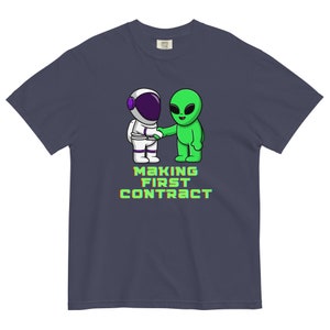 Może przedstawiać: Granatowa koszulka z kreskówkową ilustracją astronauty i kosmity podających sobie ręce. Tekst "MAKING FIRST CONTRACT" jest wyświetlany w kolorze zielonym i żółtym. Koszulka jest wykonana z miękkiego, wygodnego materiału.