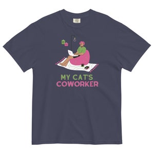 以下が含まれることがあります： 紺色のTシャツに、ピンクと緑のイラストが描かれています。イラストは、ビーズクッションに座ってラップトップで作業をしている人を表しています。イラストの下には、「MY CAT'S COWORKER」という文字がピンクと緑で書かれています。