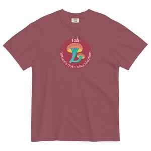 Puede incluir: Una camiseta color marrón rojizo con un gráfico circular de dos setas naranjas con tallos verdes. El gráfico tiene el texto "fall" en la parte superior y "nature's data visualization" alrededor de las setas.