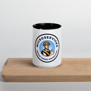 Puede incluir: Taza de cerámica blanca con borde negro. La taza presenta un logotipo circular con fondo azul y un trabajador de la construcción dando un pulgar hacia arriba. El logotipo dice "Microservices Break Down Monoliths".