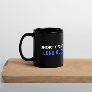 以下が含まれることがあります： 側面に白い文字で「SHORT PRINT」、青い文字で「LONG ODDS」とプリントされた黒いセラミック製のマグカップ。マグカップは標準的な円筒形で、左側に湾曲したハンドルが付いており、シンプルで機能的なデザインとなっています。