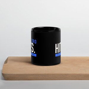 May include: A black ceramic mug featuring white and blue text. The text reads ZERO HITS. FULL CONFIDENCE. The mug has a cylindrical shape with a handle on the side and a smooth, matte finish. The design is printed clearly on the front of the mug.