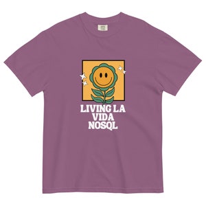 以下が含まれることがあります： 黄色の正方形の中に、緑の葉と白い星がついた笑顔の黄色い花が描かれた紫色のTシャツ。グラフィックの下には「LIVING LA VIDA NOSQL」というテキストがあります。