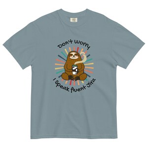 May include: A light blue t-shirt with a sloth holding a cup of coffee. The sloth is surrounded by a colorful sunburst design. The text on the shirt reads "Don't worry I speak fluent Jira".