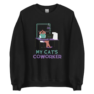 以下が含まれることがあります： 黒いスウェットシャツ。机とコンピューター、窓のある机の前にいる人のグラフィック。テキスト「MY CAT'S COWORKER」が紫とティールで下に印刷されています。スウェットシャツは柔らかい素材で作られています。