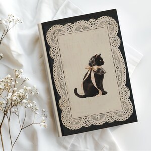 K&ouml;nnte beinhalten: Ein schwarzes Notizbuch mit festem Einband und einem cremefarbenen rechteckigen Mittelfeld, das von einer komplizierten beigen Spitzenbord&uuml;re umrahmt wird. In der Mitte ist eine Illustration einer schwarzen Katze mit Spitzenkragen und Schleife.