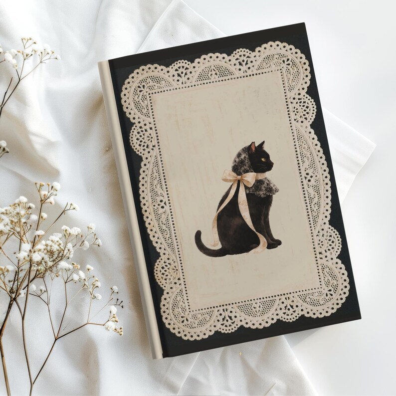 K&ouml;nnte beinhalten: Ein schwarzes Notizbuch mit festem Einband und einem cremefarbenen rechteckigen Mittelfeld, das von einer komplizierten beigen Spitzenbord&uuml;re umrahmt wird. In der Mitte ist eine Illustration einer schwarzen Katze mit Spitzenkragen und Schleife.
