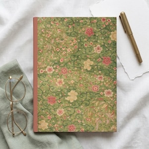May include: A rectangular notebook with a green cover featuring a repeating pattern of pink and cream-colored flowers with dark green swirling vines. A gold-toned metal pen rests on a white paper sheet next to the notebook.