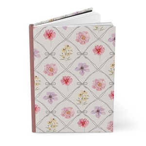 Puede incluir: Un diario de tapa dura con un estampado floral. La portada presenta un dise&ntilde;o de cuadr&iacute;cula con flores de acuarela en rosa, morado y amarillo. El diario tiene un lomo rosa y un fondo gris claro.