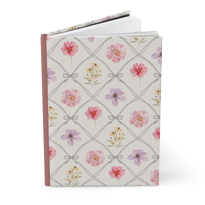 Puede incluir: Un diario de tapa dura con un estampado floral. La portada presenta un dise&ntilde;o de cuadr&iacute;cula con flores de acuarela en rosa, morado y amarillo. El diario tiene un lomo rosa y un fondo gris claro.