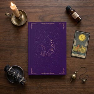 May include: A purple hardcover journal featuring a gold foil illustration of a rabbit sitting on a crescent moon surrounded by stars. The journal is accompanied by a lit candle in a brass holder, a small bottle labeled Tarot blend, a Moon tarot card, and a brass pendulum.