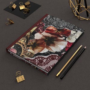 May include: A hardcover journal with a floral and lace design. The cover features large, detailed flowers in shades of brown, red, and white, with black lace accents. Gold and black pencils and gold binder clips are also visible.