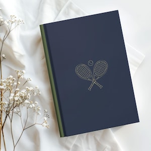 May include: A navy blue notebook with a gold tennis racket and ball design. The notebook has a green spine and is resting on a white cloth. The design features two crossed tennis rackets and a tennis ball.