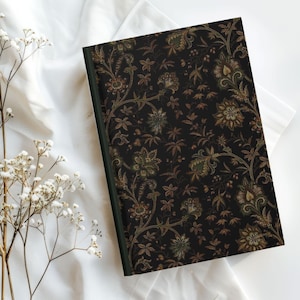 May include: A hardcover journal with a dark background and a floral pattern in shades of green and brown. The journal has a green spine and is lying on a white surface. The floral design is intricate and covers the entire cover.