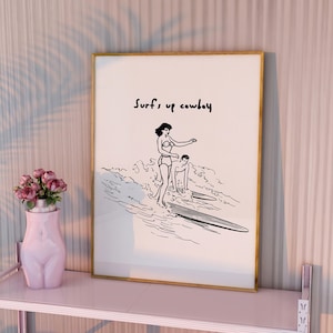 May include: A framed black and white illustration of two people surfing with the text "Surf's up cowboy" above them. The frame is gold and the illustration is on a white background. The frame is sitting on a pink shelf.