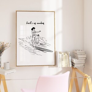 May include: Black and white line art print of two people surfing. The print has the text "Surf's up cowboy" at the top.