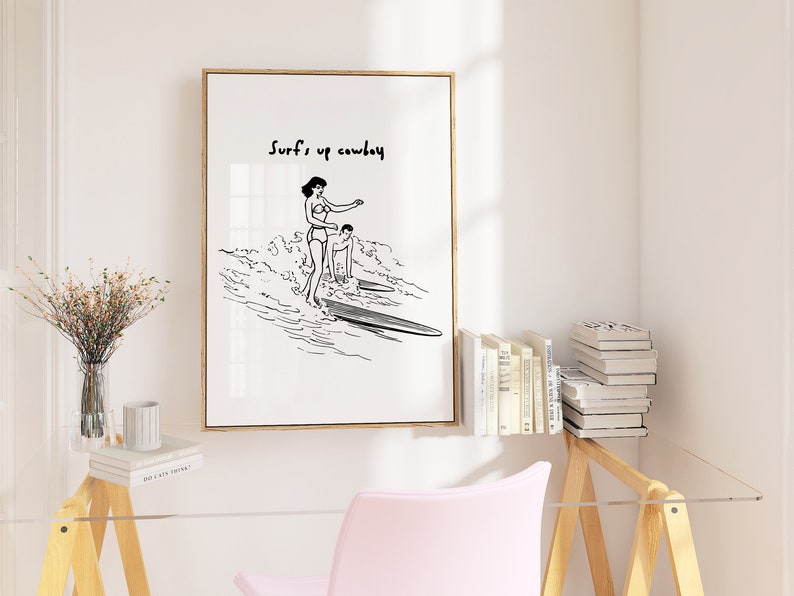 May include: Black and white line art print of two people surfing. The print has the text "Surf's up cowboy" at the top.