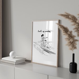 May include: Black and white line art print of two people surfing with the text "Surf's up cowboy" at the top.