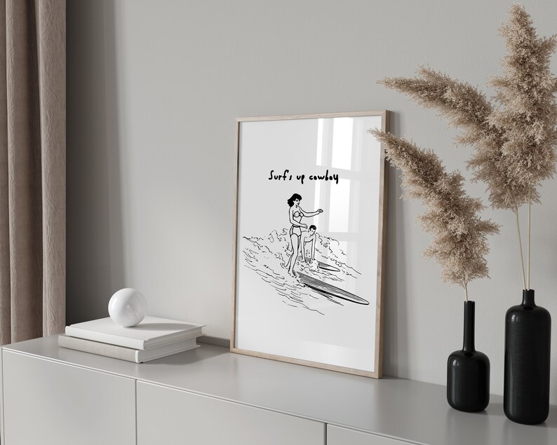May include: Black and white line art print of two people surfing with the text "Surf's up cowboy" at the top.