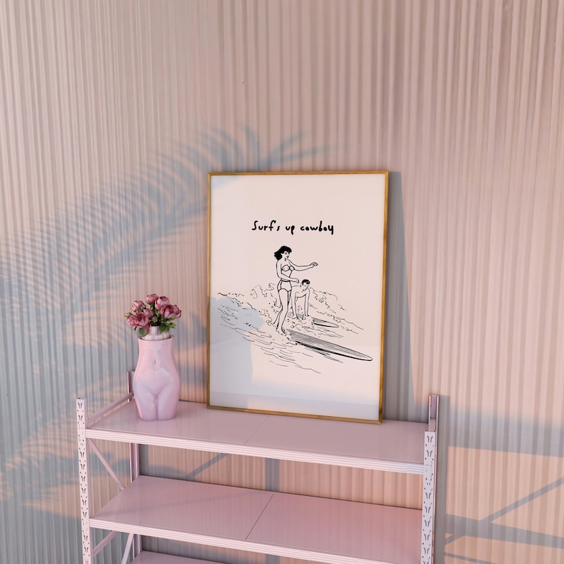 May include: A framed black and white illustration of two people surfing with the text "Surf's up cowboy" above them. The frame is gold and the illustration is on a white background. The frame is sitting on a pink shelf.