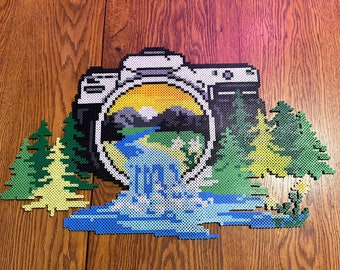 May include: A camera made of colorful beads with a landscape scene inside the viewfinder. The scene includes a blue river, green mountains, and a yellow sun.