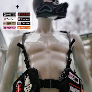May include: A black harness with patches that read "THERAPY DOG" and "EMOTIONAL SUPPORT". The harness is worn by a mannequin wearing a black and teal dog-style hood. Several stickers with the text "YOUR TEXT" are also included.