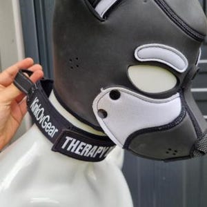 May include: A black and white pup hood with a collar that reads "KinkOvGear" and "THERAPY". The hood has a dog-like design with white accents around the eyes and muzzle. The product is displayed on a white mannequin head.