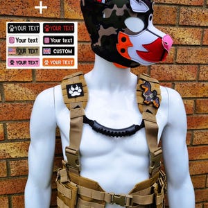 May include: A mannequin wearing a dog-themed mask with camouflage and orange accents. The mannequin also wears a tan tactical vest with multiple pockets and straps. Patches with paw prints and a dog are visible. Text on the side includes "YOUR TEXT" and "CUSTOM".