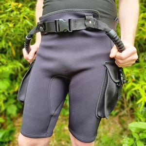 Puede incluir: Pantalones cortos de neopreno negros con cinturón incorporado y dos bolsillos laterales. El cinturón tiene una hebilla de plástico negro y dos asas. Los pantalones cortos están diseñados para actividades acuáticas.