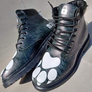 May include: A pair of black leather boots with white paw prints on the front. The boots have black laces and a black sole. The boots have a lock symbol on the top of the right boot.